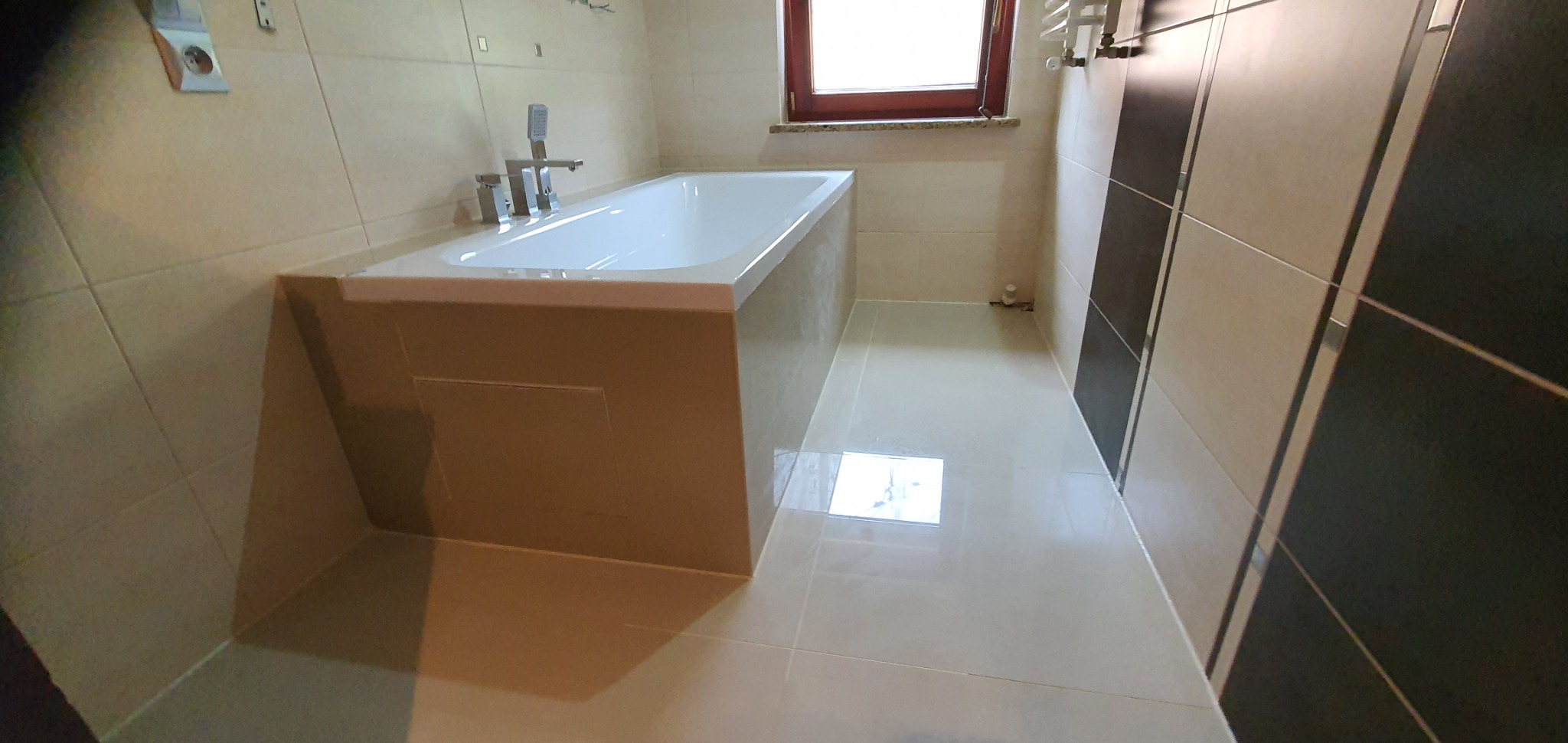 Wykończona łazienka z wanną obudowaną płytkami w odcieniach beżu, jasna podłoga, ściana z płytek beżowo-czarnych, grzejnik drabinkowy i okno z drewnianą ramą.