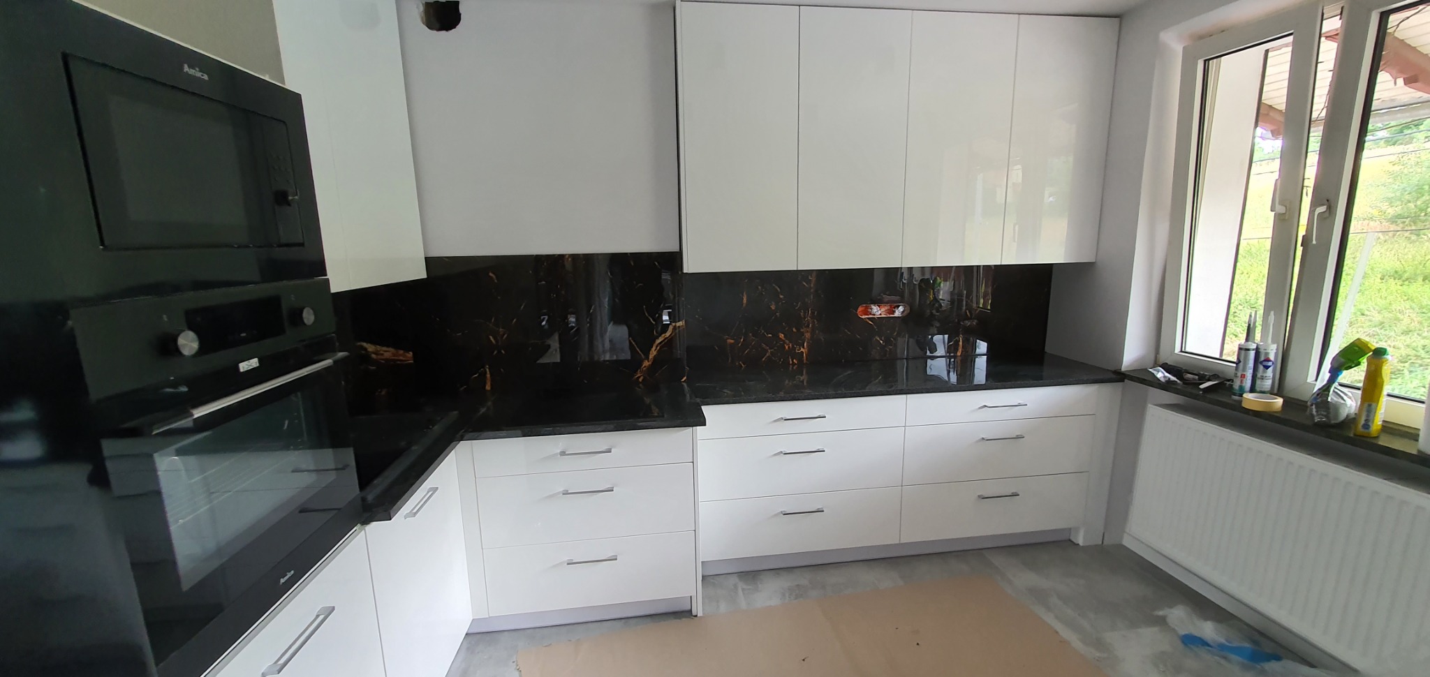 Nowoczesna kuchnia z białymi, lakierowanymi szafkami, czarnym blatem i panelem ściennym imitującym marmur, oraz czarnym piekarnikiem marki Amica w zabudowie, widok na okno z akcesoriami montażowymi.