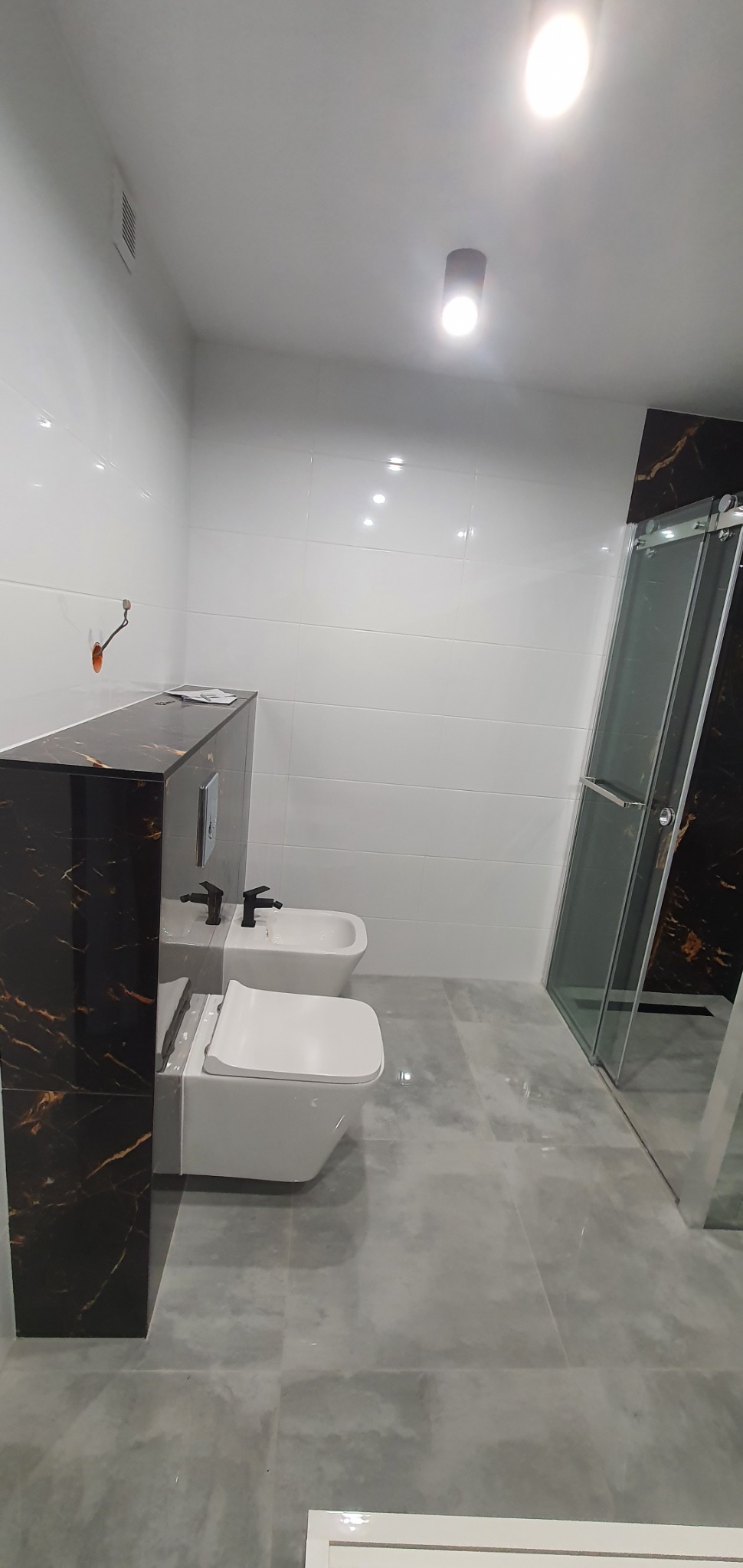 Nowoczesna łazienka po remoncie z wiszącą toaletą, bidetem z czarną armaturą, kabiną prysznicową z czarnymi akcentami i szarymi płytkami podłogowymi.