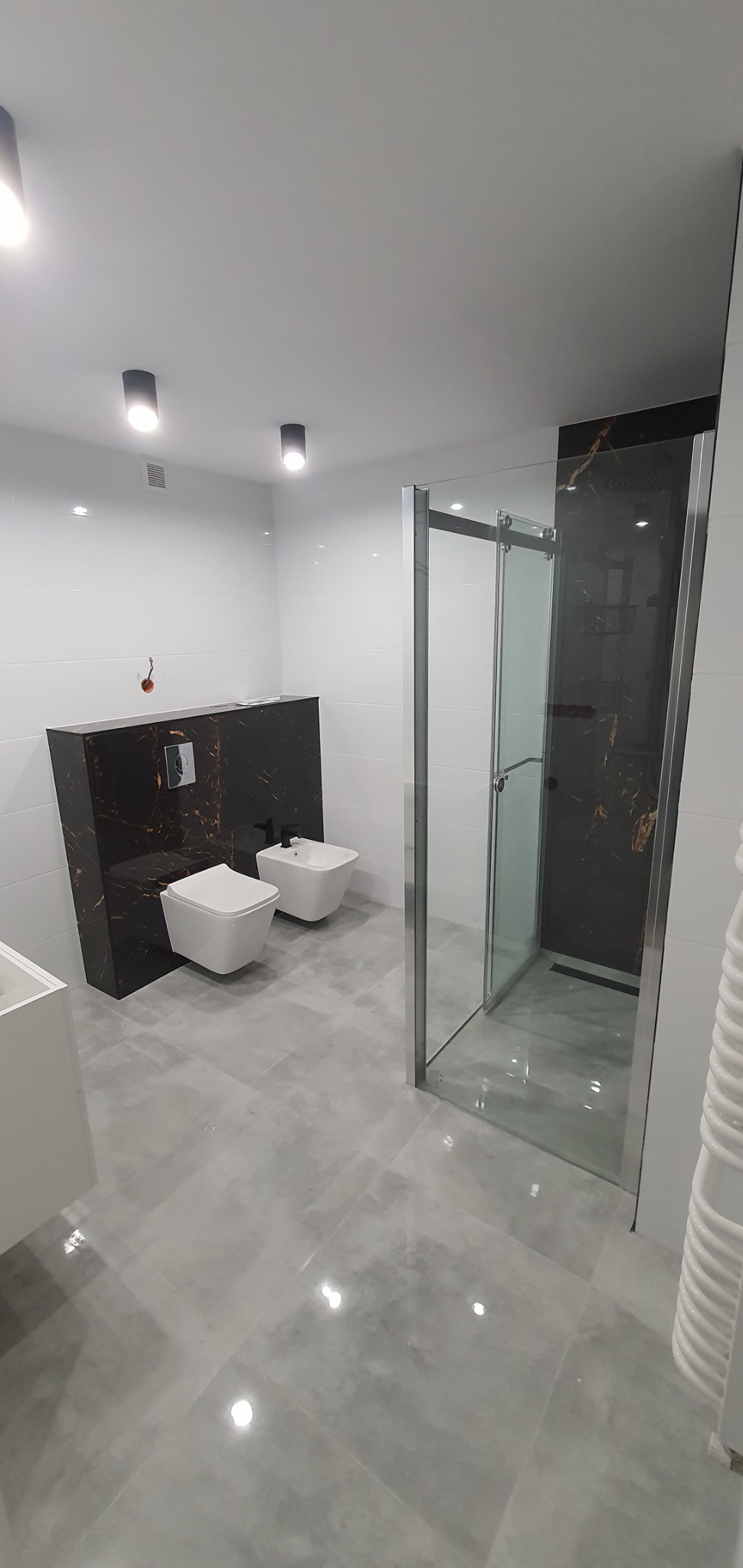 Nowoczesna łazienka z szarą, połyskującą podłogą, kabiną prysznicową ze szklanymi drzwiami, białą ceramiką sanitarną i czarnym panelem z marmurowym wzorem za toaletą i bidetem.