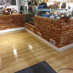 Wykończona podłoga z paneli drewnopodobnych w kawiarni, widok na ceglany bar z witryną, kratki wentylacyjne w ścianie.