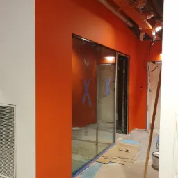 Pomarańczowa ściana świeżo po malowaniu w pomieszczeniu biurowym z przeszkloną ścianą działową zabezpieczoną taśmą, widoczne ślady farby na podłodze i kartony zabezpieczające.