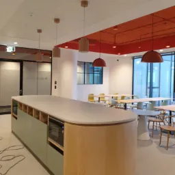 Nowoczesna przestrzeń biurowa z dużą wyspą kuchenną, stołami i krzesłami w jasnych kolorach, oświetlona wiszącymi lampami w odcieniach brązu i pomarańczy, z widokiem na budynek biurowy za oknem.