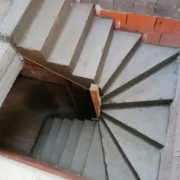 Betonowe schody spiralne w surowym stanie, widok z góry, z widoczną konstrukcją i elementami murarskimi w tle.