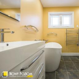 Nowy Poziom - Żółta łazienka z białą wanną wolnostojącą, umywalką nablatową i grzejnikiem drabinkowym, widoczny fragment lustra i logo firmy w rogu.