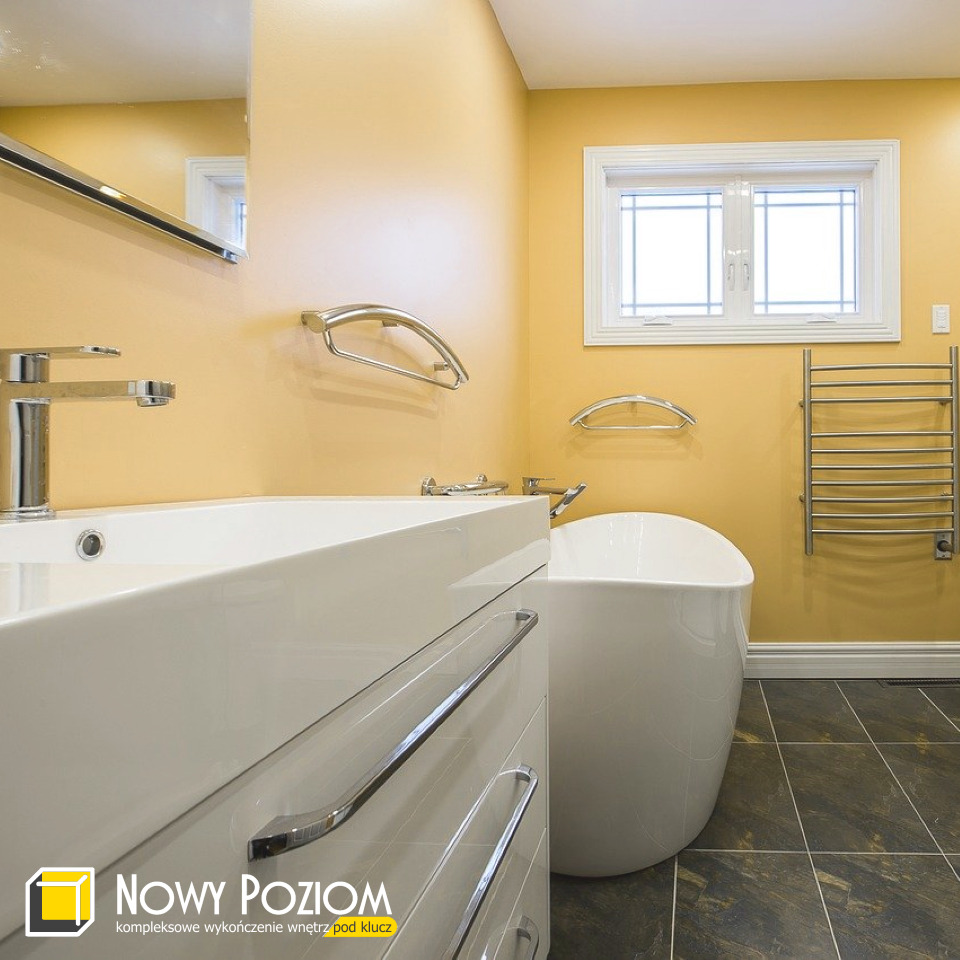 Żółta łazienka z białą wanną wolnostojącą, umywalką nablatową i grzejnikiem drabinkowym, widoczny fragment lustra i logo firmy w rogu.