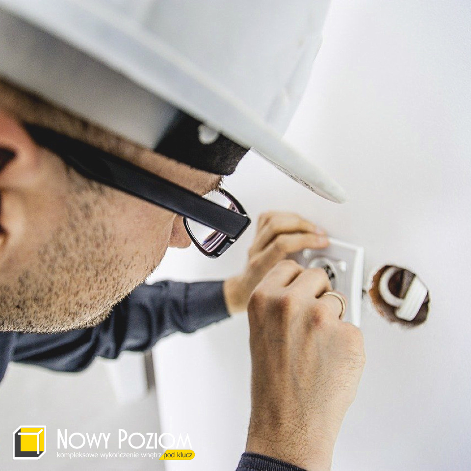 Elektryk w okularach i kasku montuje gniazdko elektryczne w białej ścianie, widoczne przewody, logo firmy w rogu.