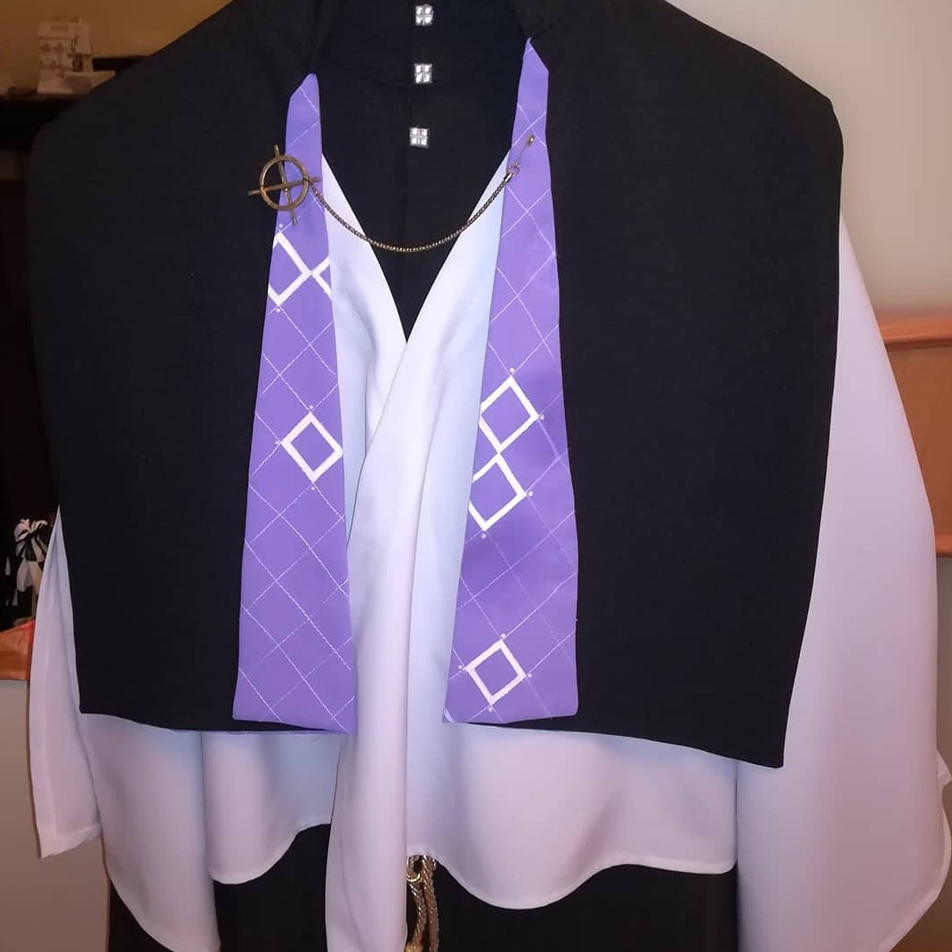 Czarno-biała szata z fioletowym, romboidalnym elementem dekoracyjnym przypiętym łańcuszkiem i ozdobną broszką, widoczne detale wykończenia.