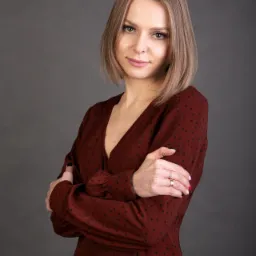 Portret kobiety z blond włosami, ubranej w bordową sukienkę w czarne kropki, na szarym tle. Ręce skrzyżowane, widoczne pierścionki na palcach.