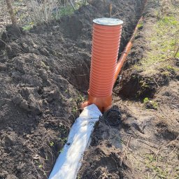 Zielono Mi - Pomarańczowa rura drenażowa w wykopie ziemnym, zabezpieczona białą geowłókniną. W tle widoczny pracownik przy kolejnym elemencie systemu.