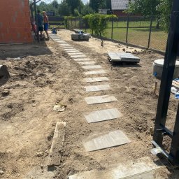 Zielono Mi - Układanie ścieżki z płyt chodnikowych na gruncie, widoczne narzędzia i materiały budowlane. W tle budynek w trakcie budowy i ogrodzenie posesji.
