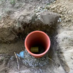 Pomarańczowa rura drenażowa w wykopie, otoczona kamieniami. Wewnątrz widoczna mniejsza, żółta rura. Tło stanowi ziemia i trawa. Specyficzne ułożenie rur.