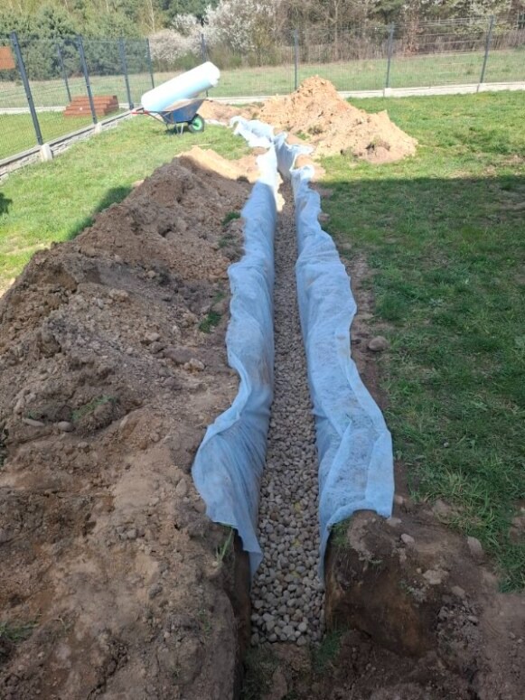 Wykop wypełniony żwirem, zabezpieczony geowłókniną, prawdopodobnie drenaż. W tle taczka z rolką materiału i pryzma ziemi.