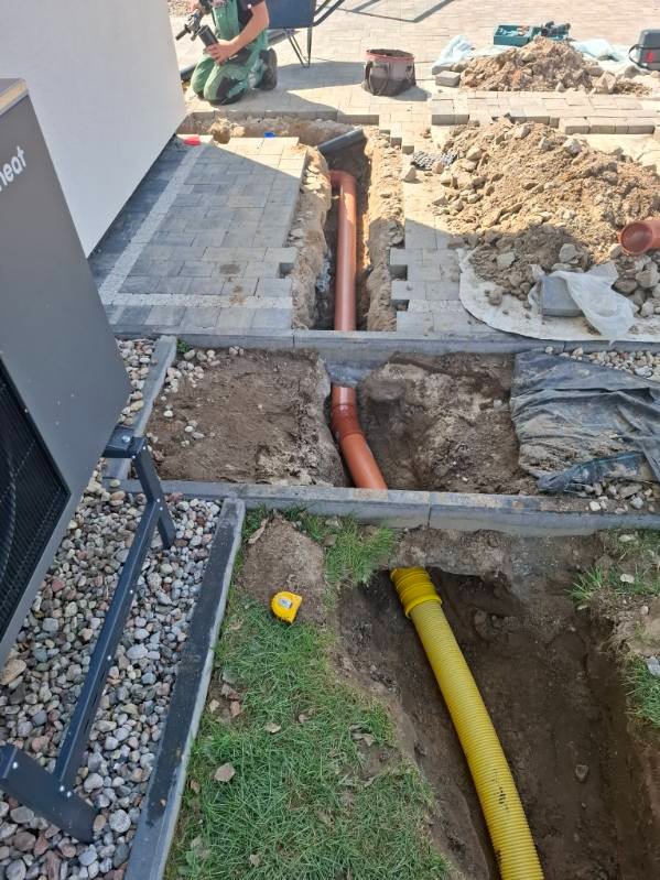 Wykop z ułożonymi rurami drenażowymi w ziemi, widoczne różne typy rur (żółta karbowana i pomarańczowa gładka). Częściowo ułożona kostka brukowa. Prace ziemne.