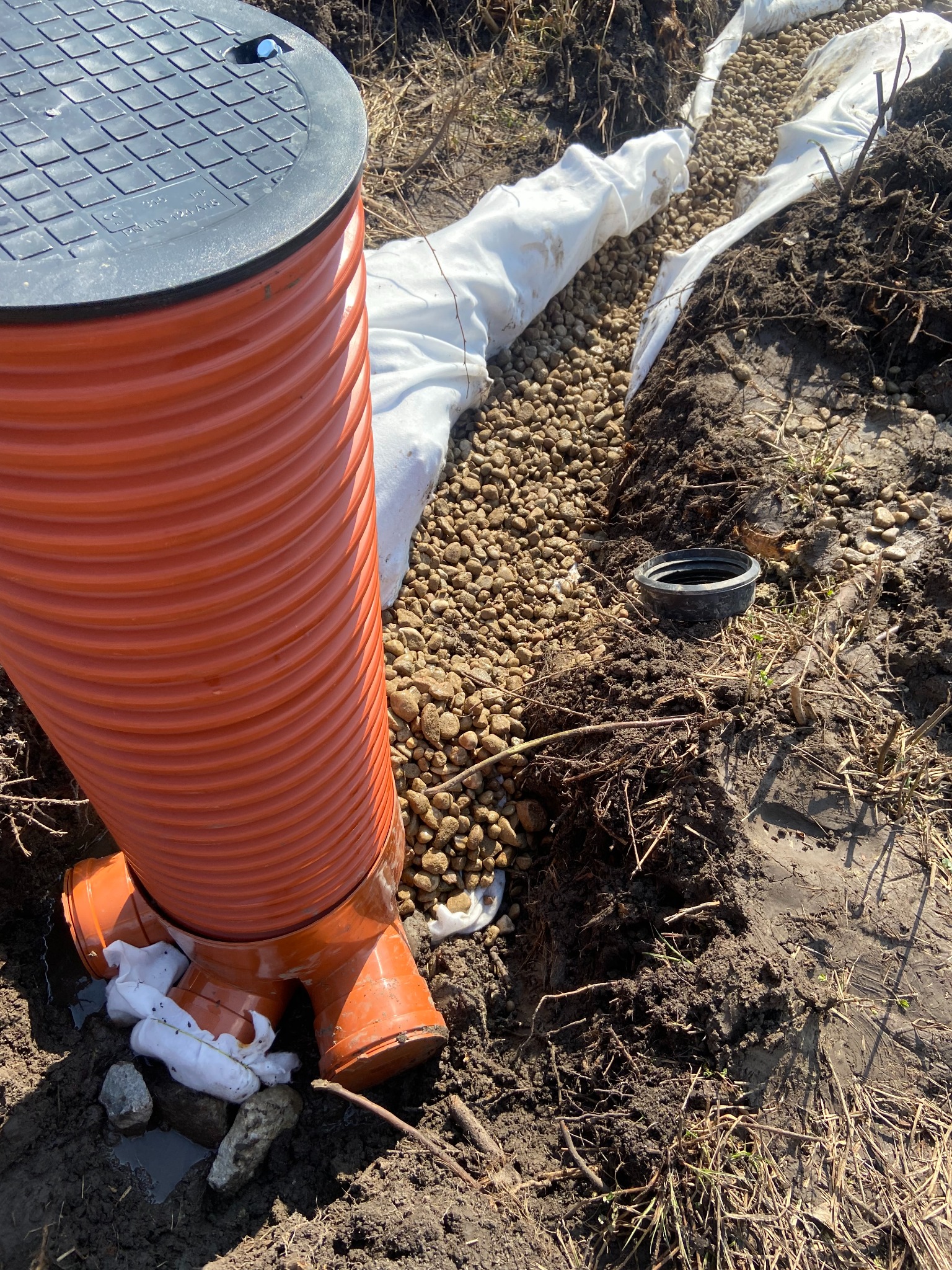 Pomarańczowa rura drenażowa z czarną pokrywą studzienki, otoczona kamieniami i białą geowłókniną w wykopie. Widoczne elementy instalacji drenażowej.