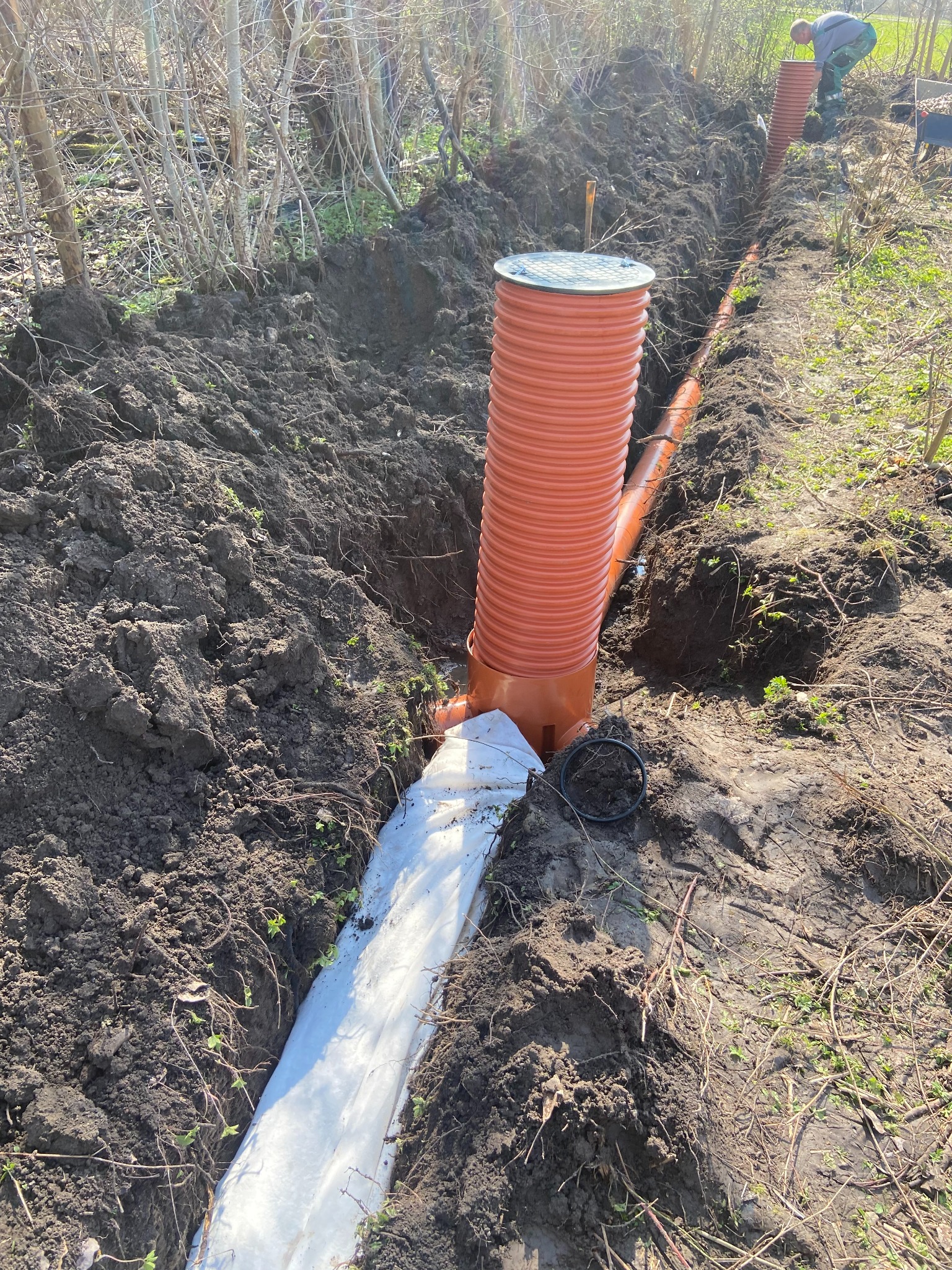 Pomarańczowa rura drenażowa w wykopie ziemnym, zabezpieczona białą geowłókniną. W tle widoczny pracownik przy kolejnym elemencie systemu.