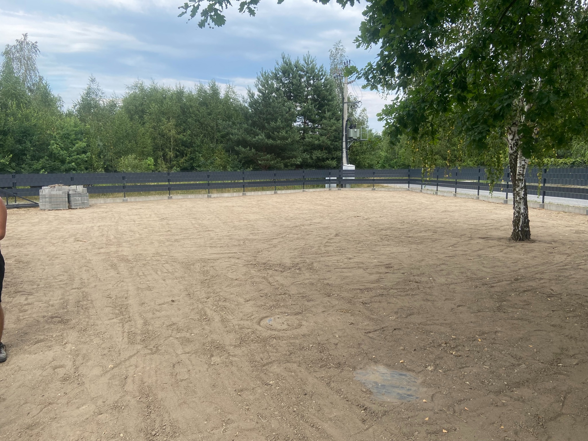 Wyrównany teren pod budowę ogrodzony czarnym płotem, z widocznym drzewem i stertą betonowych bloczków. W tle las i słup energetyczny.