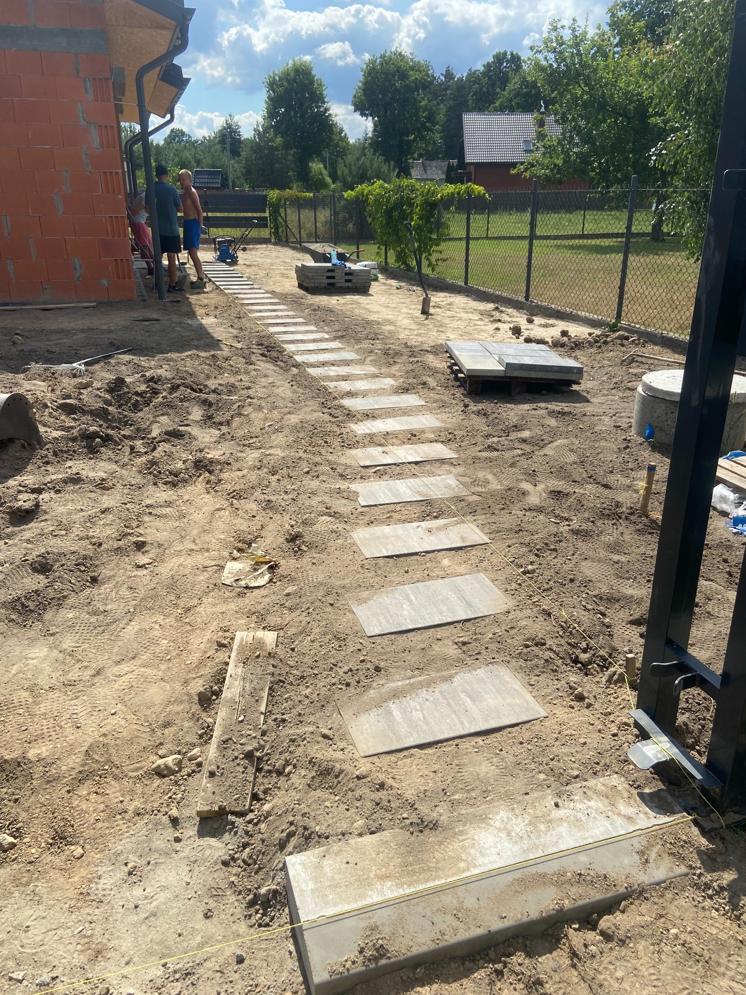 Układanie ścieżki z płyt chodnikowych na gruncie, widoczne narzędzia i materiały budowlane. W tle budynek w trakcie budowy i ogrodzenie posesji.