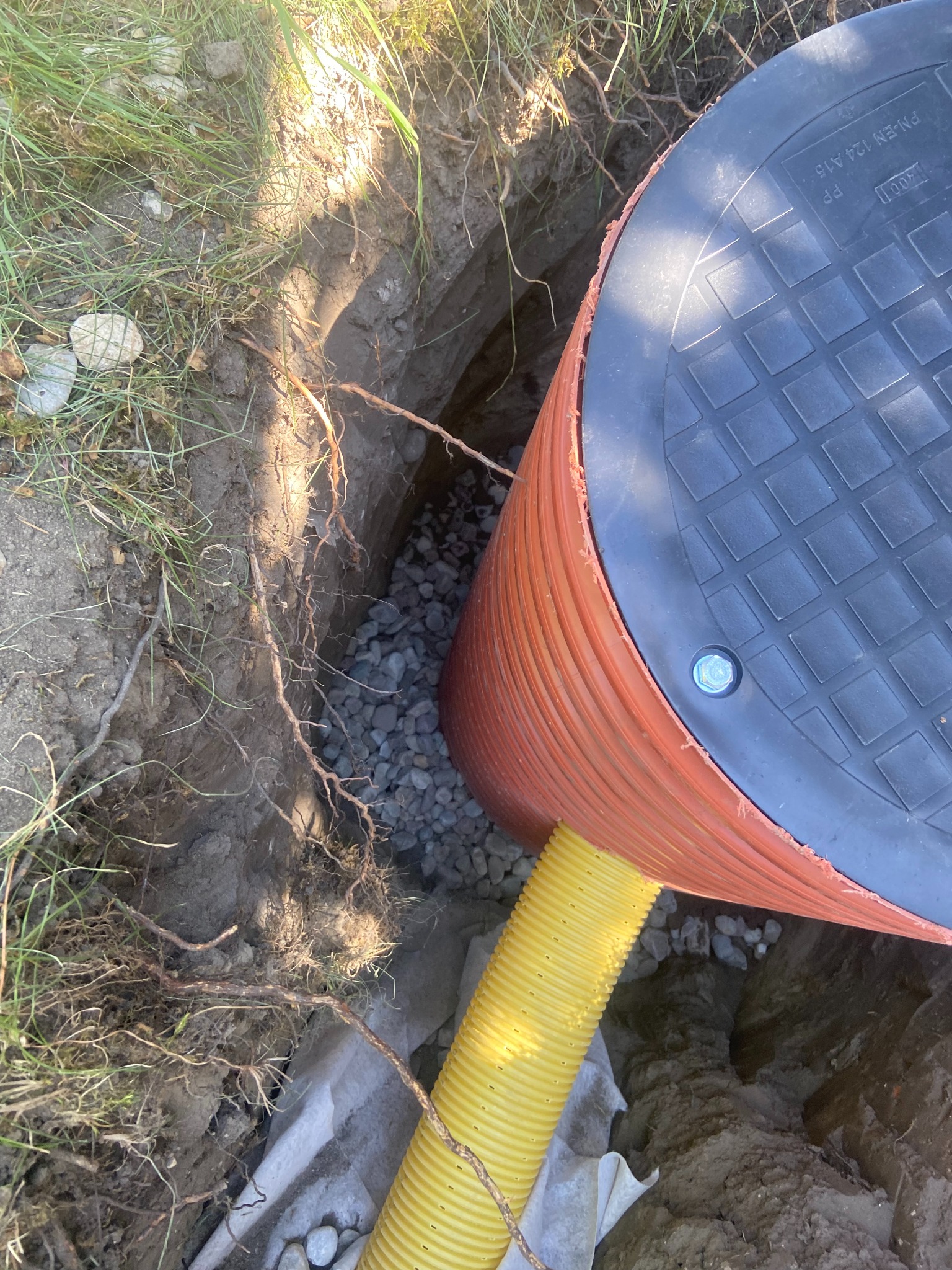 Pomarańczowa rura z czarnym deklem studni w wykopie, otoczona kamieniami i żółtą rurą drenażową. Widoczne korzenie i trawa na krawędzi wykopu.