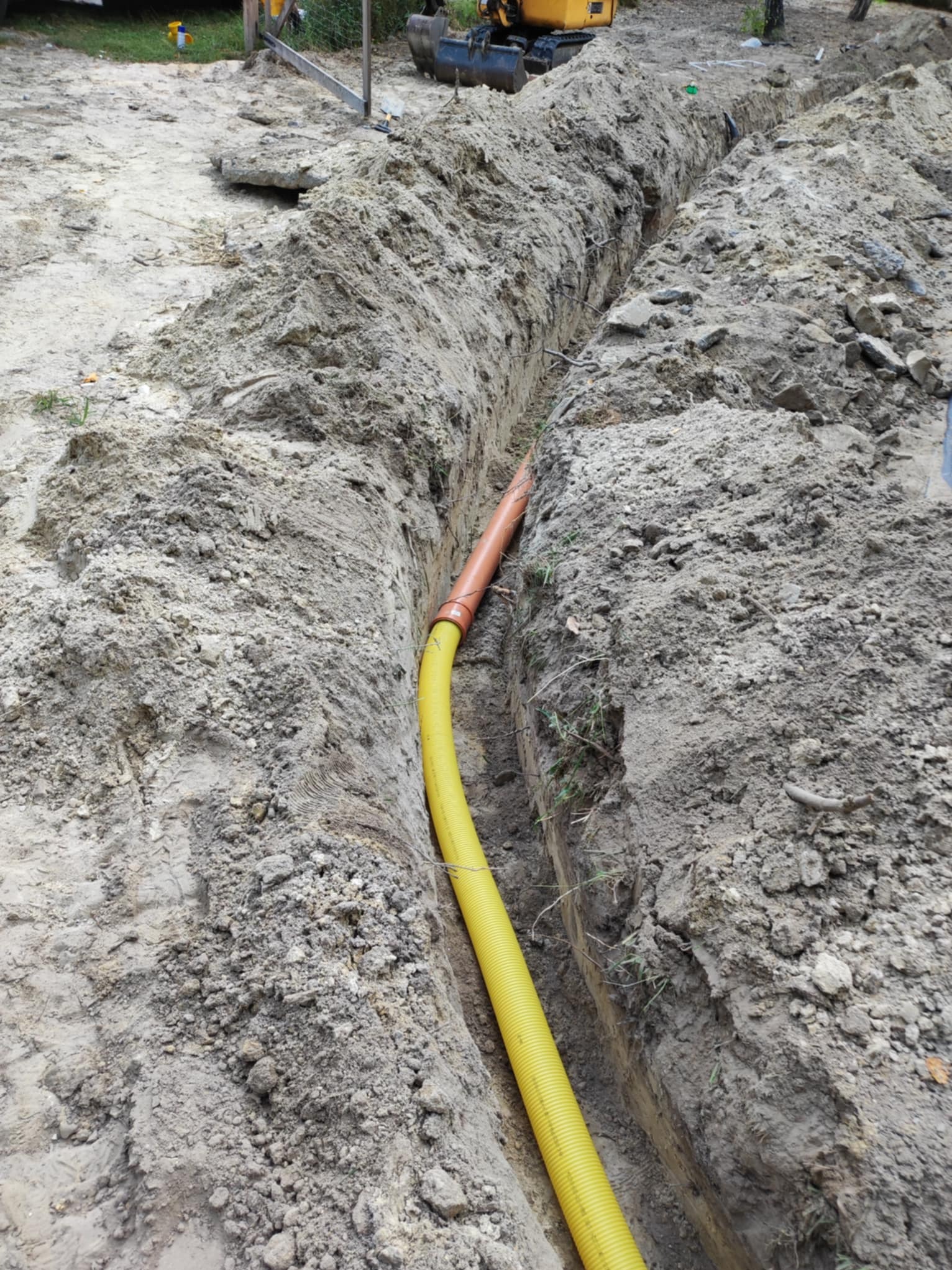 Żółta rura karbowana i pomarańczowa rura gładka w wykopie ziemnym. W tle widoczny sprzęt budowlany. Realizacja robót ziemnych.