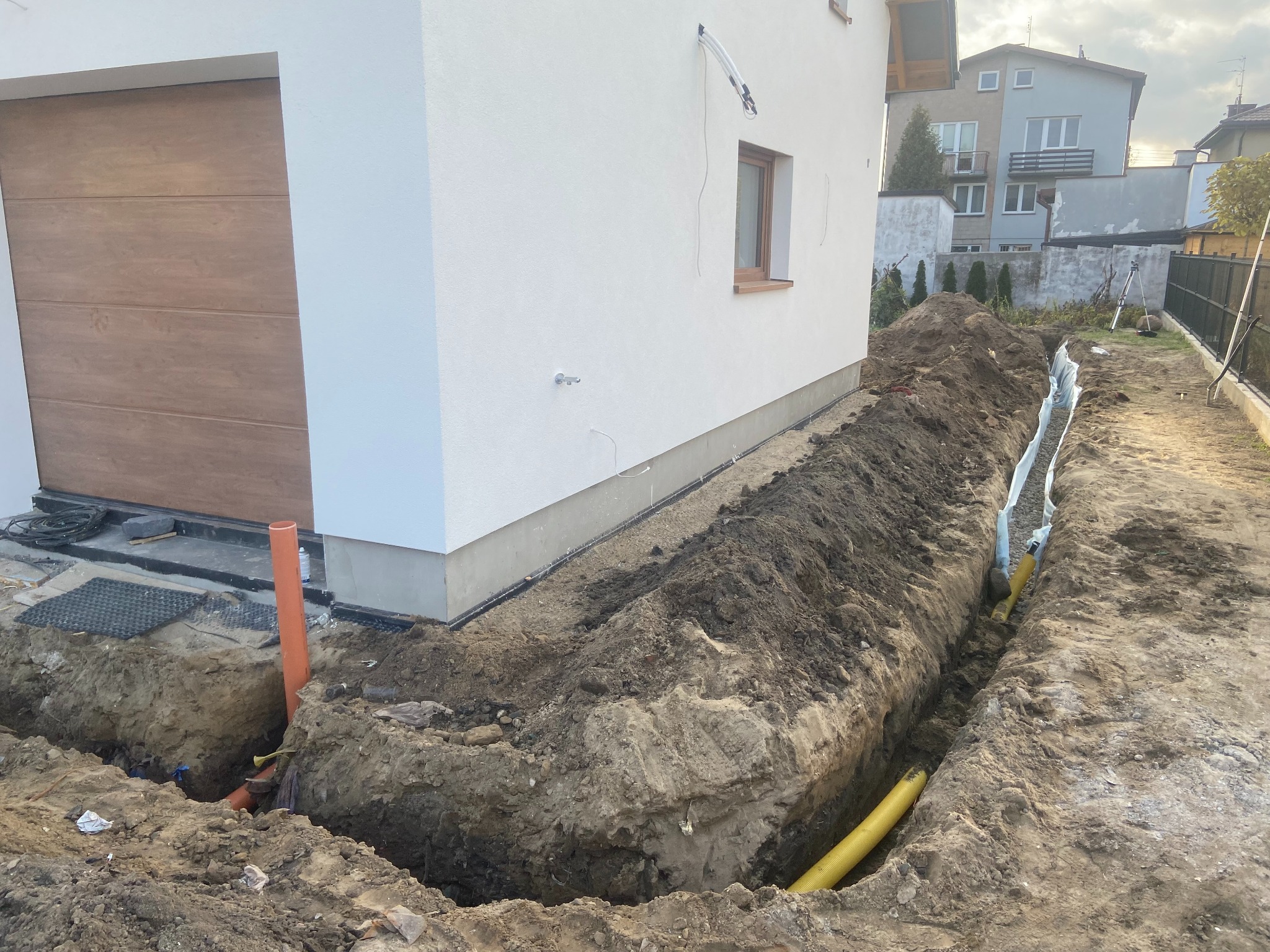 Wykop pod fundamentem domu z widocznymi rurami drenażowymi i instalacyjnymi. W tle budynek mieszkalny i garaż z drewnianą bramą.