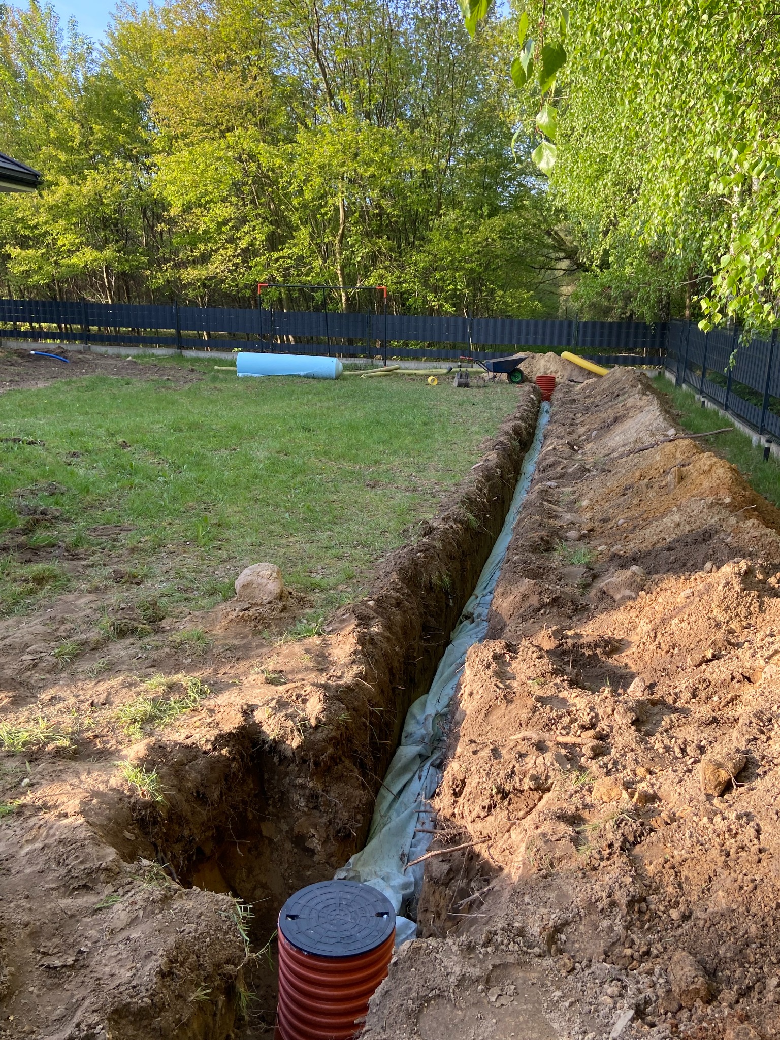 Wykopany rów w ogrodzie, wyłożony geowłókniną, z widoczną rurą drenarską z tworzywa i studzienką rewizyjną. W tle trawnik, ogrodzenie i drzewa.