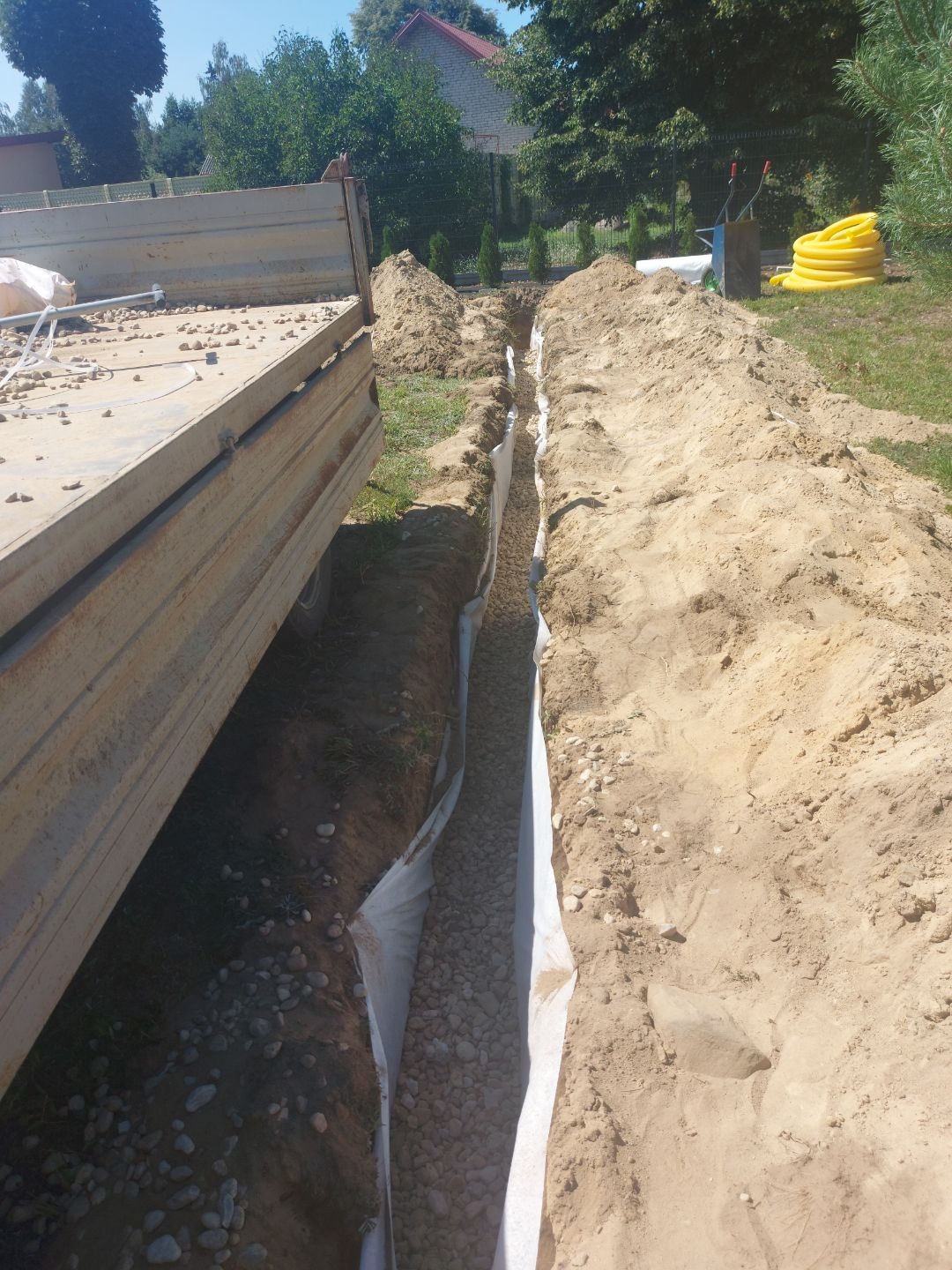 Wykopany rów z geowłókniną i kamieniami, obok zaparkowana ciężarówka z kamieniem, w tle widoczne drzewa i budynek. Prace ziemne.