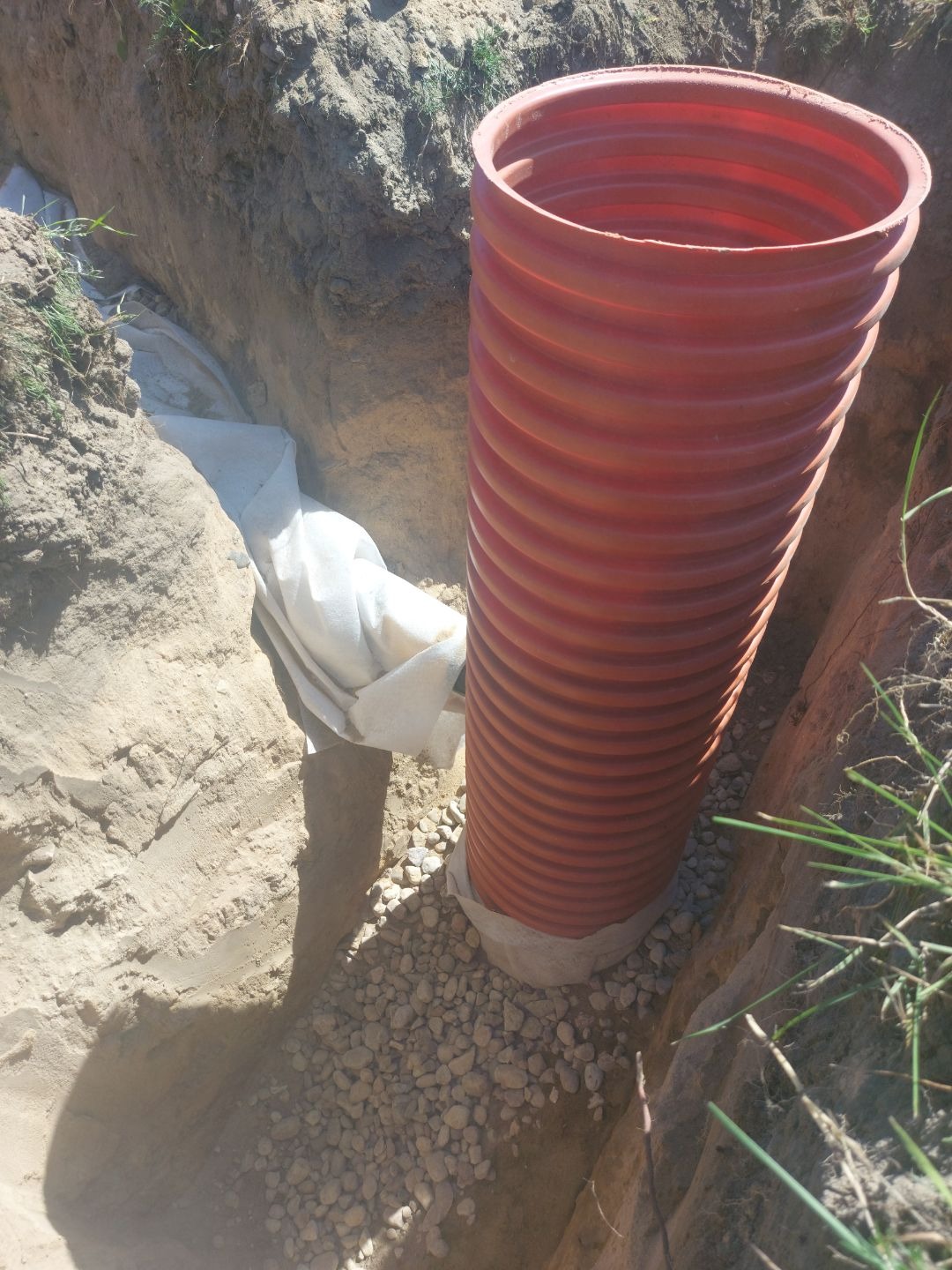 Instalacja pionowa rura drenarska z geowłókniną w wykopie wypełnionym żwirem. Rura karbowana, pomarańczowa, wystaje ponad poziom gruntu. Drenaż.