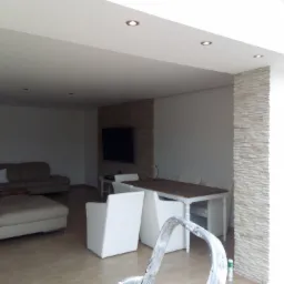 Jasne wnętrze salonu z fragmentem ściany wykończonej dekoracyjnym kamieniem, widoczne meble wypoczynkowe i stół z krzesłami, na pierwszym planie fragment drabiny malarskiej.