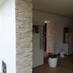 Fragment ściany z dekoracyjną okładziną kamienną w jasnych odcieniach, oddzielający przestrzeń w nowoczesnym wnętrzu. Widoczny biały mebel i roślina doniczkowa w tle.