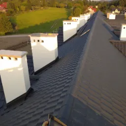 Nowy, czarny dach z ceramicznej dachówki, z rzędem białych kominów na tle zielonych wzgórz w słoneczny dzień.