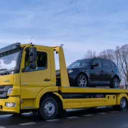 Żółta laweta Mercedes-Benz z czarnym SUV-em na platformie, widziana w Tychach na tle błękitnego nieba i drzew bez liści.