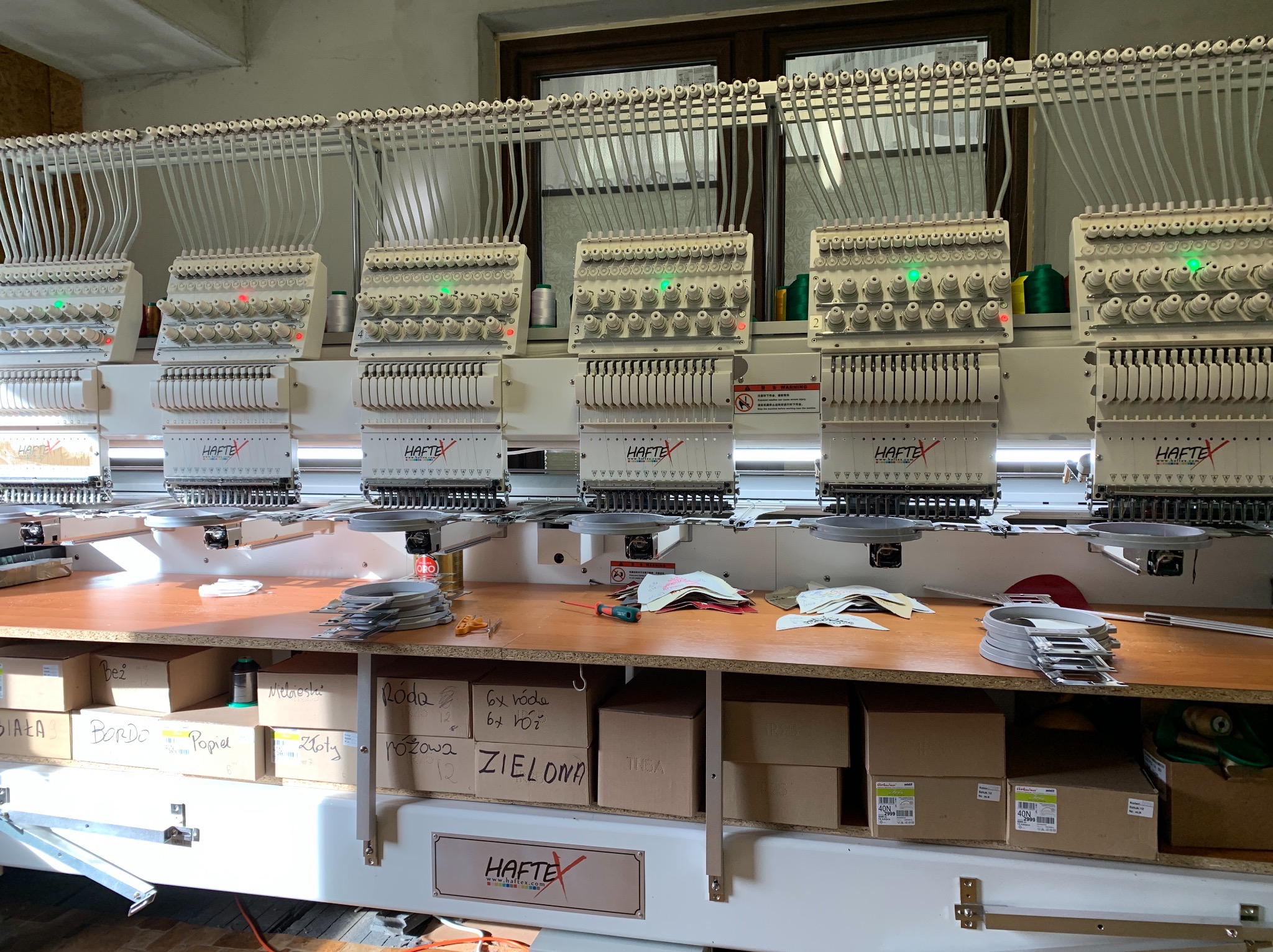 Wielogłowicowa maszyna hafciarska z nawiniętymi nićmi różnej barwy, widoczne pudełka z opisami kolorów nici: biały, bordo, popiel, złoty, zielona, róda. Na blacie leżą tamborki i narzędzia.