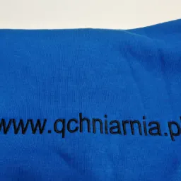 Niebieska tkanina z wyhaftowanym czarnym napisem 'www.qchniarnia.pl'.