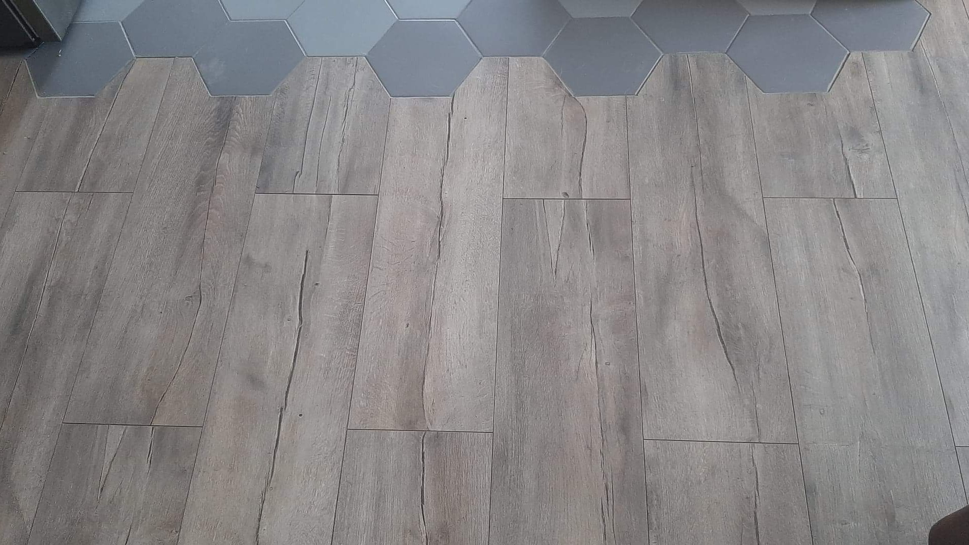 Ułożona podłoga z paneli imitujących drewno w odcieniach szarości, z fragmentem szarej, heksagonalnej płytki ceramicznej przy ścianie.