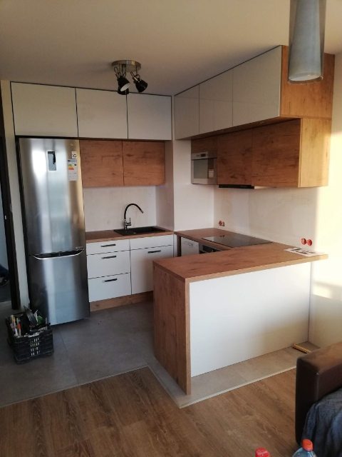 Nowoczesna kuchnia z białymi i drewnopodobnymi szafkami, srebrną lodówką i czarnym zlewem, oświetlona trzema reflektorami sufitowymi. Na podłodze szare płytki przechodzą w panele imitujące drewno.