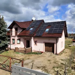 Dom jednorodzinny z panelami słonecznymi na dachu z czerwonej dachówki, widok z lekkiej góry na posesję z ogrodzeniem w trakcie budowy i drzewem iglastym po lewej stronie.