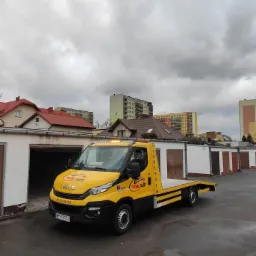 Żółto-czarna laweta marki Iveco z napisem MOBILGUM na tle rzędu garaży i bloków mieszkalnych pod pochmurnym niebem.