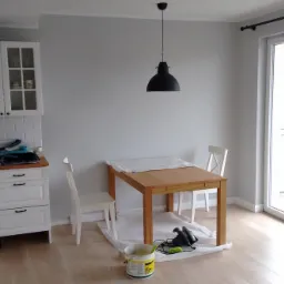 Jasne wnętrze z drewnianym stołem zabezpieczonym folią malarską, przygotowanym do malowania ścian w odcieniach szarości, widoczny kubeł z farbą i szlifierka pod stołem.