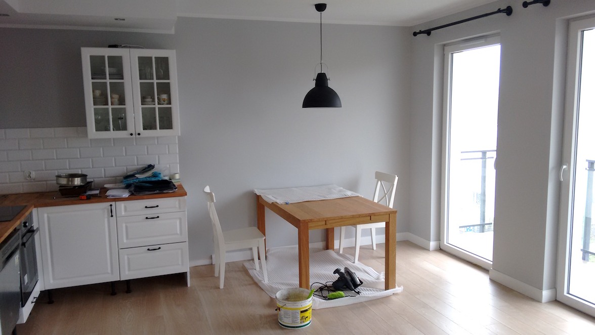 Jasne wnętrze z drewnianym stołem zabezpieczonym folią malarską, przygotowanym do malowania ścian w odcieniach szarości, widoczny kubeł z farbą i szlifierka pod stołem.