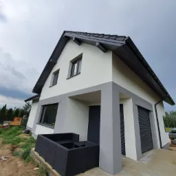 Nowoczesny dom jednorodzinny z szarą stolarką okienną i elementami elewacji, widok z dołu na tle pochmurnego nieba, z widocznym fragmentem tarasu i mebli ogrodowych.