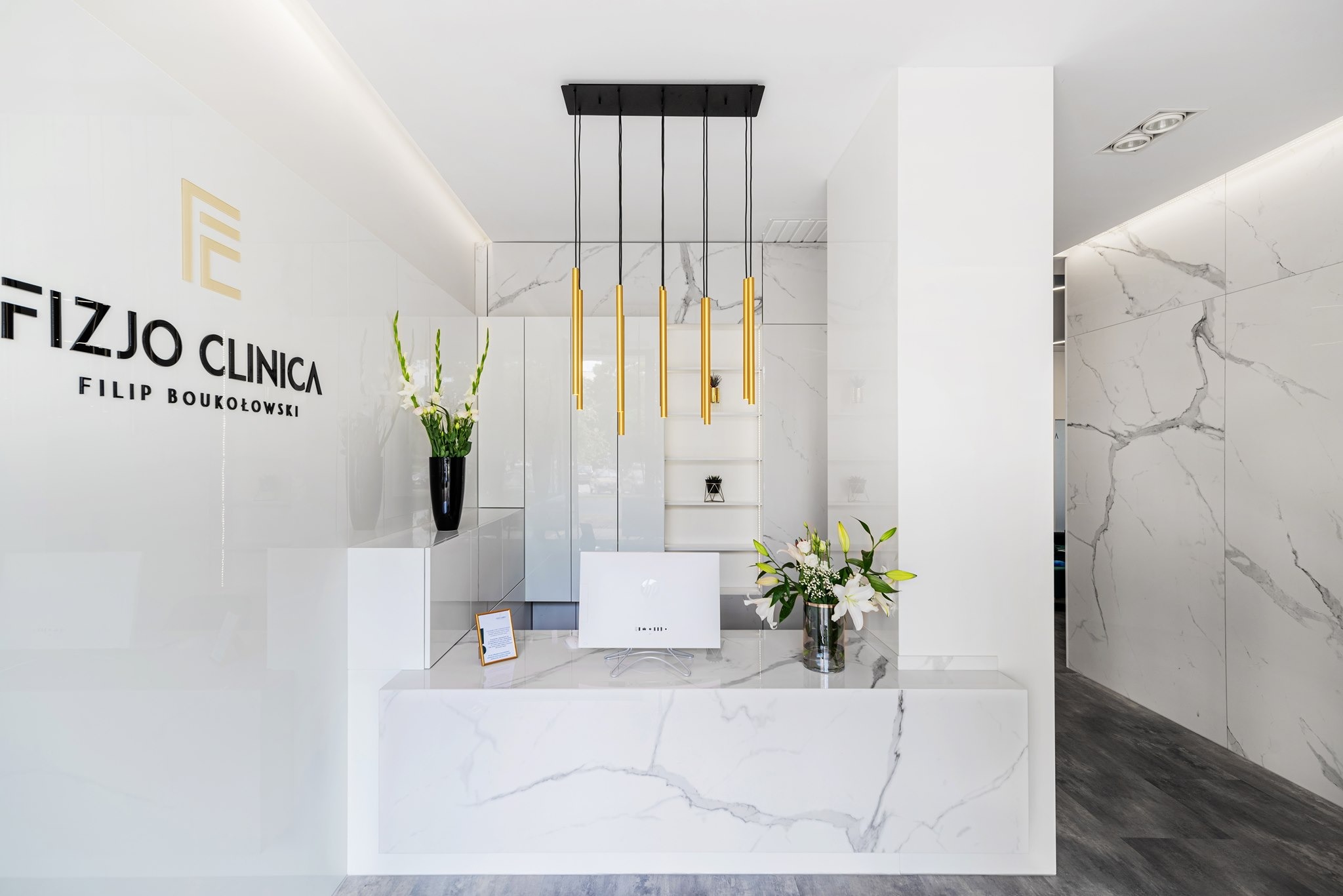 Nowoczesne wnętrze recepcji kliniki w Leoncinie, charakteryzujące się białymi ścianami z efektem marmuru, marmurowym blatem recepcyjnym i złotymi lampami wiszącymi.