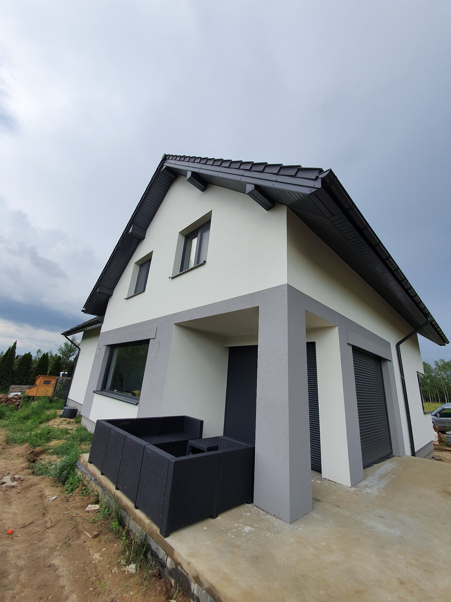 Nowoczesny dom jednorodzinny z szarą stolarką okienną i elementami elewacji, widok z dołu na tle pochmurnego nieba, z widocznym fragmentem tarasu i mebli ogrodowych.