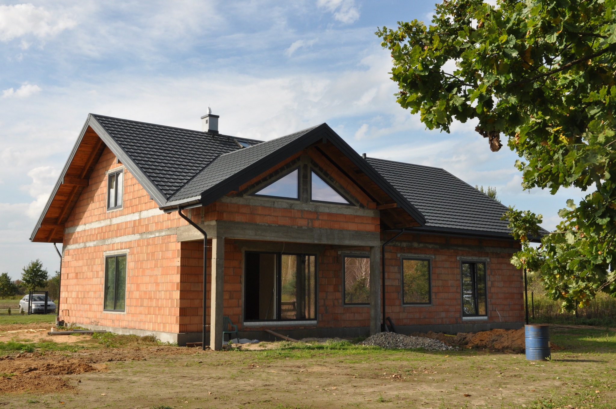 Surowy budynek jednorodzinny z czerwonej cegły z szarą dachówką, widoczne przygotowane otwory okienne i drzwiowe, otoczony zielenią i błękitnym niebem.