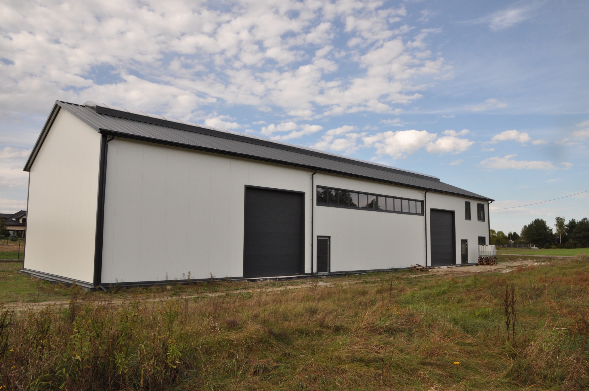 Nowoczesny, biały budynek magazynowy z czarnym dachem i bramami garażowymi, otoczony polną roślinnością, widok z zewnątrz w słoneczny dzień.