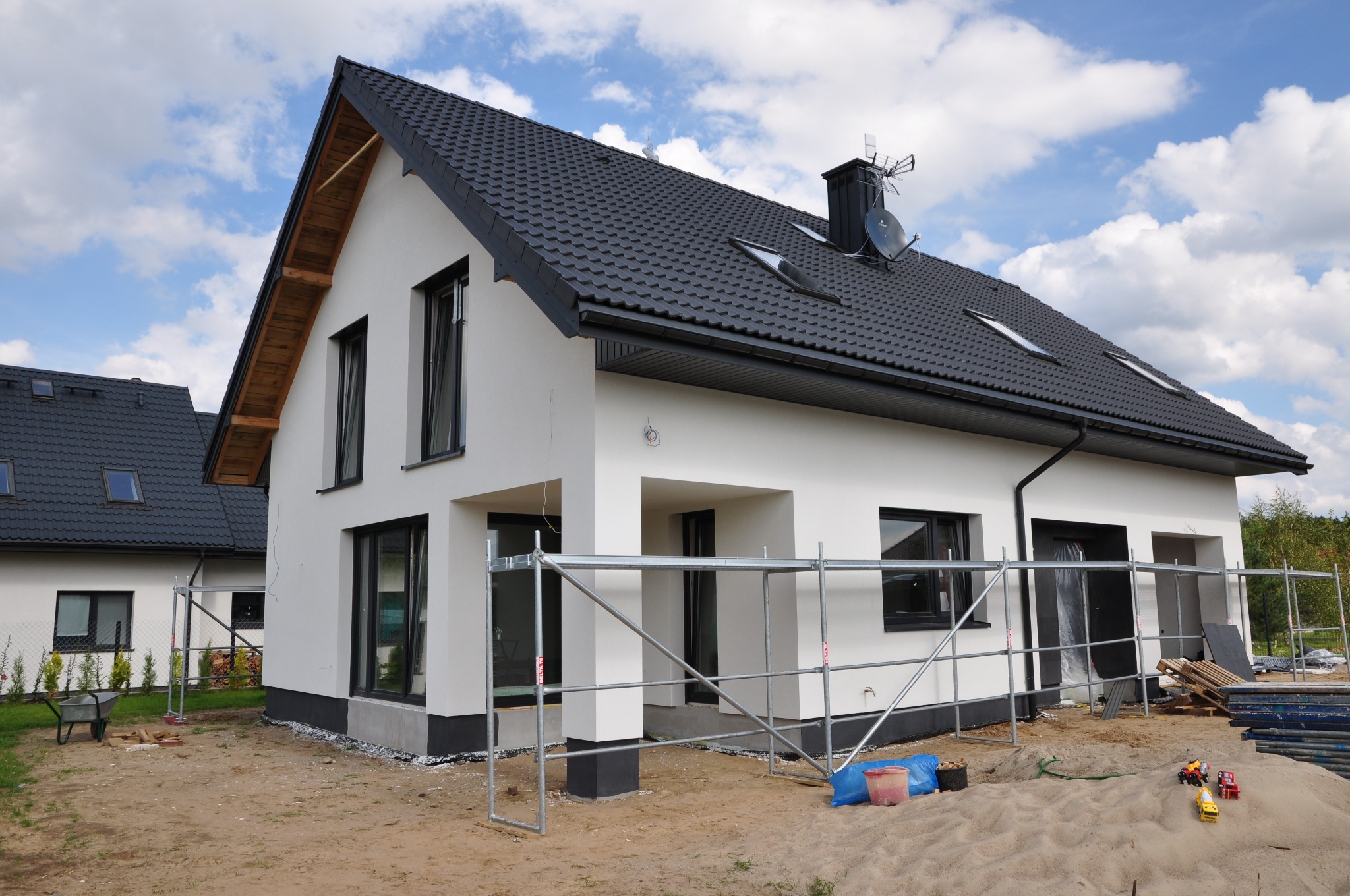 Nowoczesny, dwukondygnacyjny dom z ciemną dachówką i białymi ścianami zewnętrznymi, widoczne rusztowania i materiały budowlane przed budynkiem, sugerujące prace wykończeniowe.
