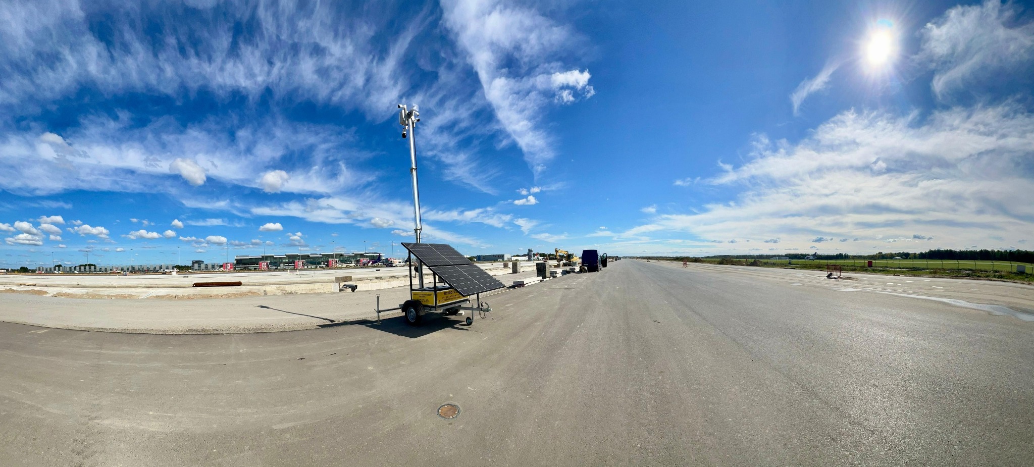 Mobilny system monitoringu z kamerami i panelem solarnym na placu budowy lotniska, błękitne niebo z chmurami w tle. Szeroki kadr, perspektywa.