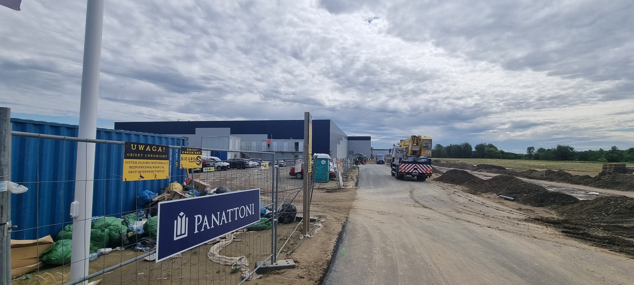 Teren budowy z widocznym magazynem w tle, ogrodzony siatką, z tablicami informującymi o ochronie obiektu, oraz logo firmy Panattoni. Na drodze wjazdowej stoi ciężarówka z dźwigiem.