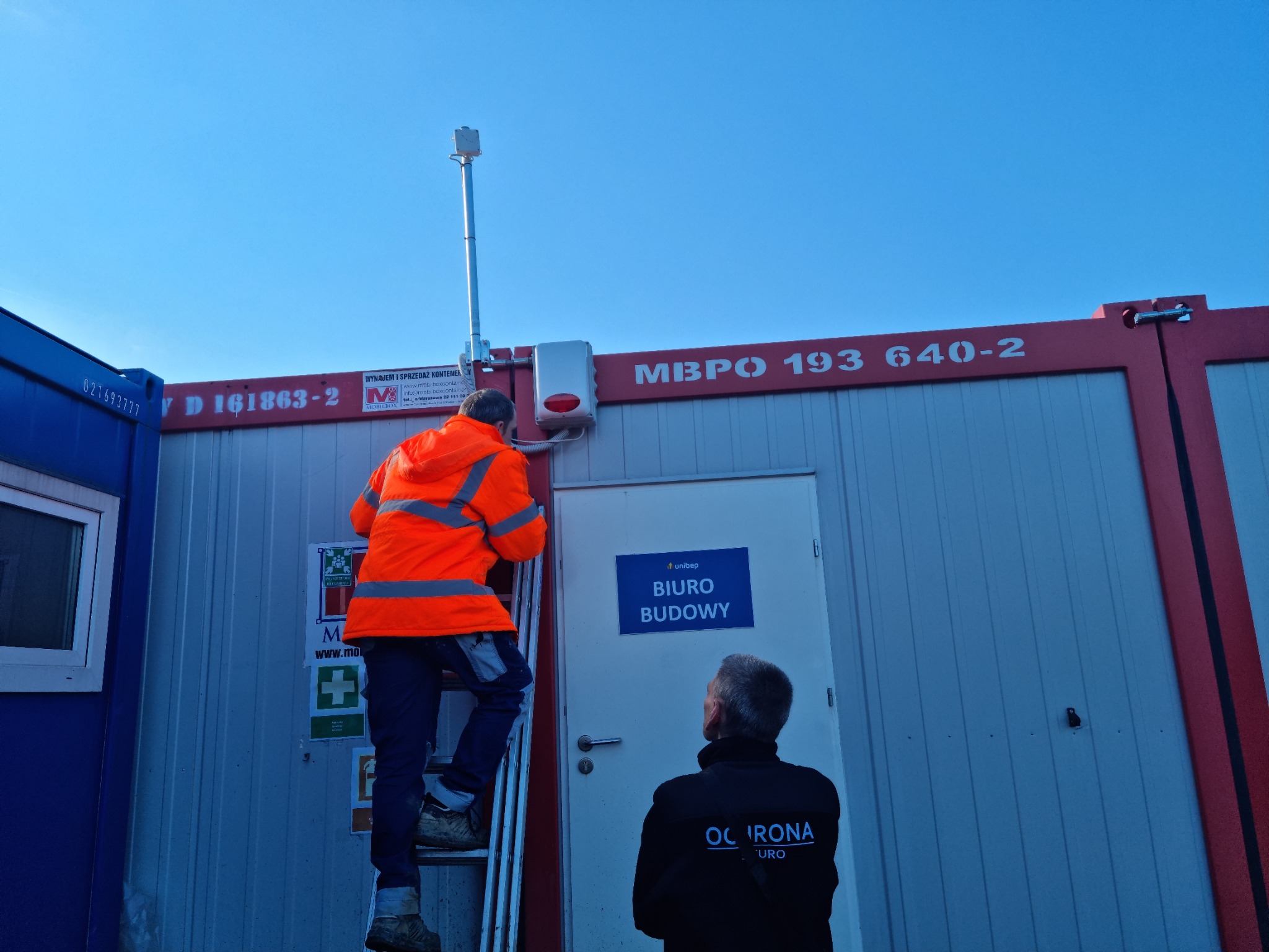 Instalator w pomarańczowej kurtce na drabinie montuje czujnik na kontenerze biurowym budowy, obok stoi pracownik ochrony w czarnej bluzie z napisem OCHRONA.