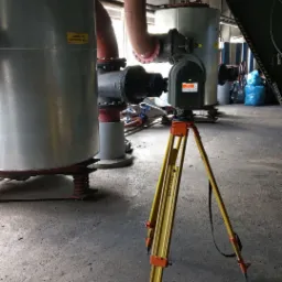 Skaner 3D na żółtym statywie Wild GST20 w hali przemysłowej, w tle zbiorniki i rury instalacji przemysłowej.